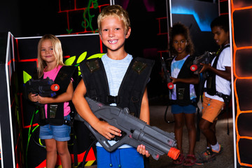 Positive teen boy standing with laser pistol in dark lasertag room during game with friends