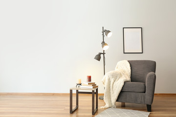 Comfortable armchair, table and lamp near white wall