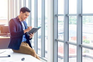 Fototapeta premium A businessman wearing a suit and a long-sleeve shirt inside and sitting smart poses in a modern office. In his hand have a pen and a notebook in action reading or writing something.