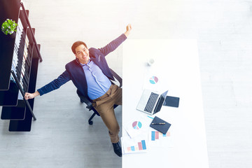 Top view of young businessman sitting in a modern office. He has a feel happy and smile about the result of business profits is positive. On his table have a computer laptop tablet pen paper graph 