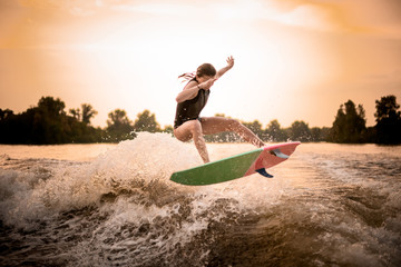 Skinny girl in black swimsuit jumping on the wakeboard on the river on the wave in the sunset