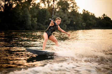 Sporty girl riding on the wakeboard on the river in the sunset holding a rope of the motorboat