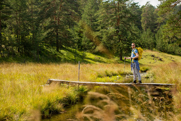 Young man with backpack crosses a mountain stream on a sunny day