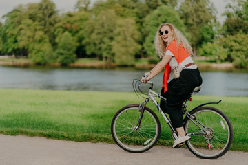 Outdoor shot of happy blonde curly woman wears sunglasses, dressed in casual active wears, rides on bicycle near lake and green trees in countryside, spends free time outside, enjoys favourite hobby