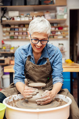 Senior female potter working on pottery wheel while sitting  in her workshop