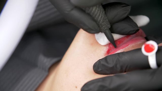 Cute brunette lying on the couch on the procedure of permanent lip makeup. Tattoo artist applies a special pigment with the pen.  Beauty industry Close up.