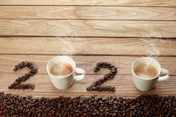Top view of coffee beans and hot fresh coffee in a white cup with foam and smoke with text 2020 Happy New Year Concept. on wooden table background.