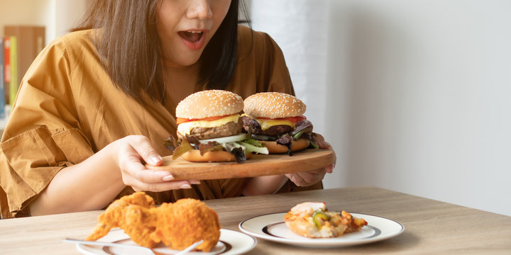 Hungry overweight woman holding hamburger on wooden plate, Fried chicken and Pizza on table .Concept of binge eating disorder (BED).