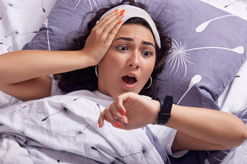 Oversleeping concept. Young woman missed ringing alarm clock, be late, looking with horror at clock...