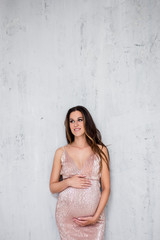 Fototapeta na wymiar beautiful pregnant woman in shiny evening dress posing over gray wall background with copy space