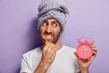 Close up shot of handsome male with stubble, wakes up in morning, holds alarm clock showing time,...