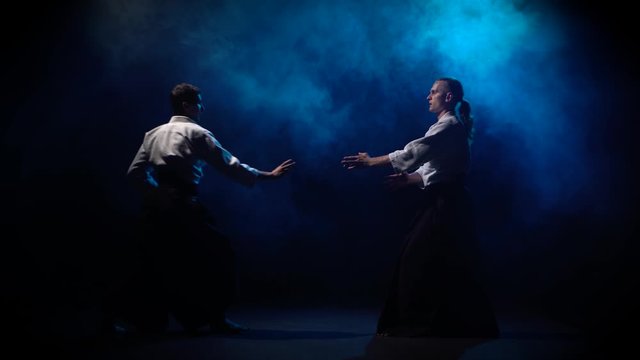 Two fighters participants of the training in special clothes of aikido hakama work out the methods of single combat on blue smoke background.