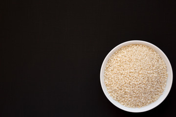 Organic Arborio rice in a white bowl on a black surface, top view. Flat lay, overhead, from above. Copy space.