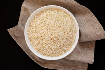 Organic Arborio rice in a white bowl on a black background, overhead view. Flat lay, top view, from above. Close-up.