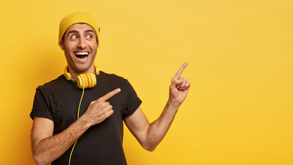 Horizontal shot of happy European man points aside with two index fingers, demonstrates awesome promo, dressed in stylish balck and yellow wear, wears modern headphones around neck for listening song