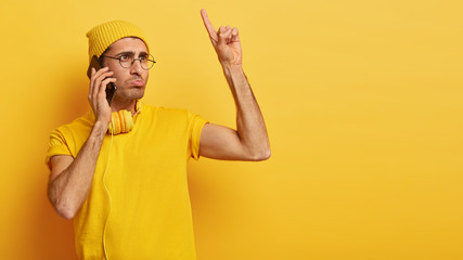 Sad displeased man has telephone conversation, raises index finger up, hears bad news, wears yellow hat and t shirt, gets idea in mind how to improve situation, stands indoor, copy space for your text