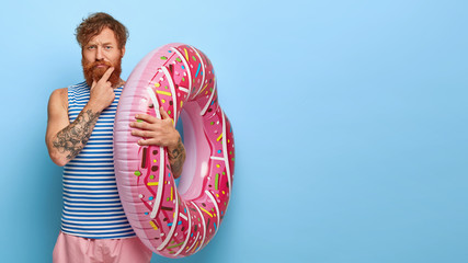 Photo of serious bearded adult ginger man holds big inflated swimring, dressed in summer wear, has...