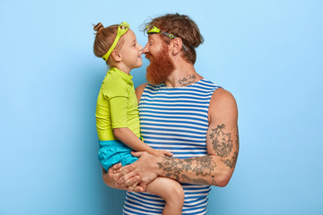 Photo of bearded redhead young man carries little daughter, touch noses and have happy expressions, wear goggles, come in swimming pool for spending day actively, express love to each other.