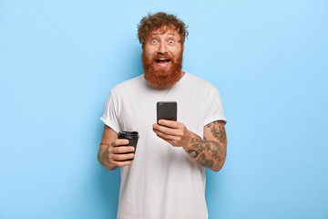 Smiling bearded emotional man has red hair, holds mobile phone, shares great news with friend,...