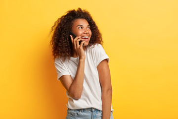 Studio shot of beautiful satisfied young African American woman enjoys pleasant conversation, holds mobile phone near ear, looks aside, wears casual white t shirt, poses against yellow wall.