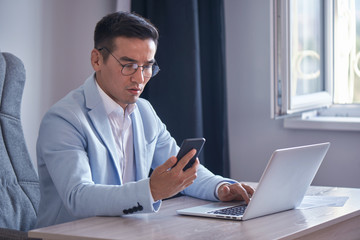 Asian Kazakh businessman in a suit and glasses with a laptop computer in the office speaks by mobile phone, solves business issues, negotiates a deal.