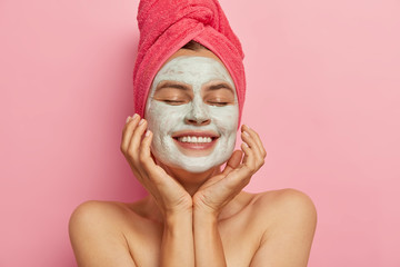 Photo of satisfied woman improves her skin condition, wears facial mask, touches cheeks, shows white perfect teeth, has towel for drying hair, makes daily beauty routine, isolated on pink wall