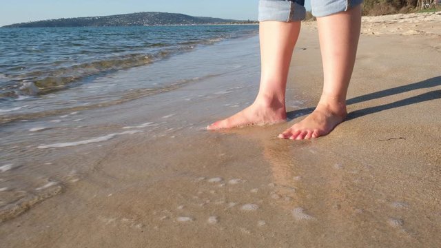 Caucasian woman feet standing on the beach washed by coming waves at sunset - low angle static shot