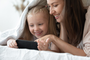 Mother and daughter lying in bed under blanket with smartphone