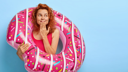 Photo of lovely cheerful woman covers mouth with palm, looks aside, imagines something pleasant, wears red swimsuit, stands with inflated donut, isolated against blue background, has good rest