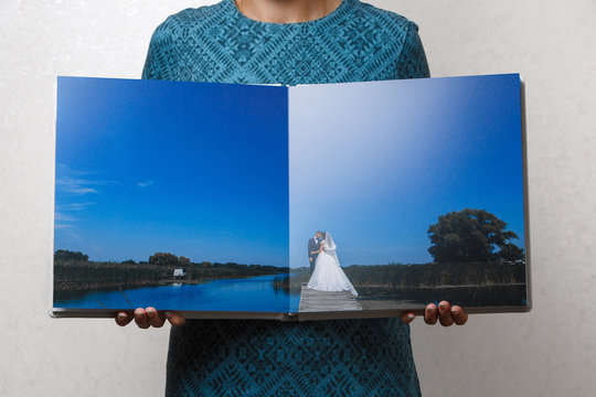 a woman holds open family photobook the person looks at the photo book sample  beige photo album  wedding photoalbum with  fabric cover.female hands holding square open photo album.