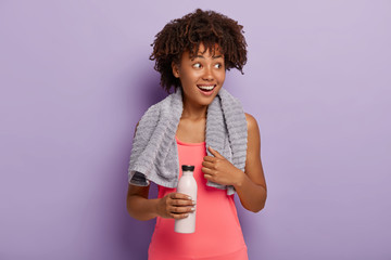 Photo of delighted dark skinned sportswoman has Afro haircut, looks aside with smile, dressed in pink top, carries bottle, drinks water while feels thirsty during training, excercises indoor.