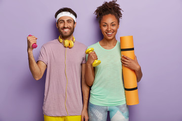 People, exercising and sport concept. Happy Caucasian man and dark skinned woman raise dumbbells, carry fitness mat, have toothy smiles, dressed in casual wear, stand indoor against purple background