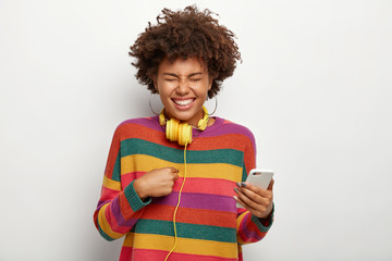 Half length shot of overjoyed cheerful woman points at herself, holds mobile phone, expresses pleasant emotions, wears earrings, colored jumper, has headphones around neck, isolated on white wall