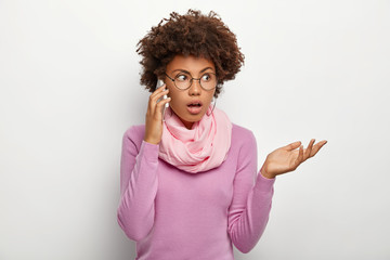 Puzzled female with Afro hairstyle, has conversation with colleague or friend, tries to solve problem via smartphone, focused aside, wears optical glasses, purple casual jumper and scarf around neck