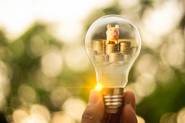 Hand holding a light bulb and stack of coins with piggy bank inside for saving money concept....