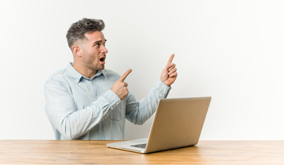 Young handsome man working with his laptop pointing with forefingers to a copy space, expressing excitement and desire.