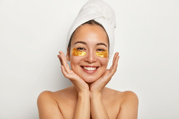 Beauty portrait of happy Asian girl keeps palms near face, looks positively at camera, shows white perfect teeth, enjoys spa procedures, stands with wrapped towel on head against white studio wall