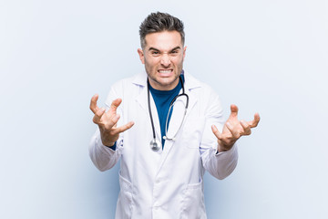 Young handsome doctor man upset screaming with tense hands.