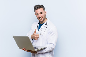 Caucasian doctor man holding a laptop smiling and raising thumb up