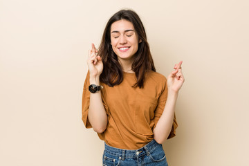 Young brunette woman against a beige background crossing fingers for having luck