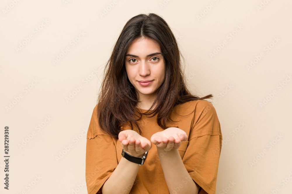 Wall mural Young brunette woman against a beige background holding something with palms, offering to camera.