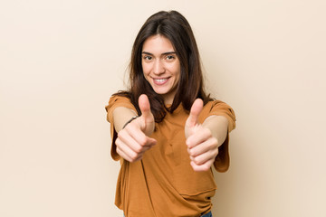 Young brunette woman against a beige background with thumbs ups, cheers about something, support...