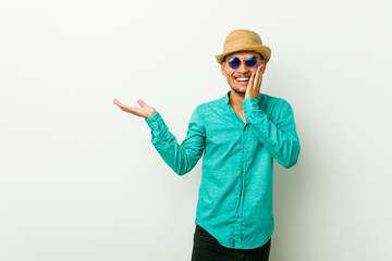 Young hispanic man wearing a summer clothes holds copy space on a palm, keep hand over cheek. Amazed and delighted.