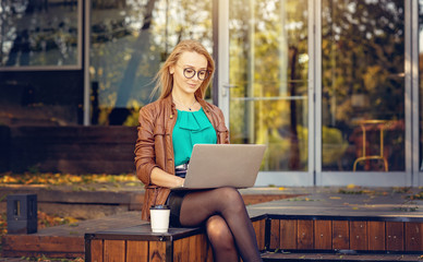 Woman working on laptop outdoors autumn in front of the entrance of a closed summer restaurant - Young woman entrepreneur working outdoors surrounded by golden autumn leaves - End of season concept