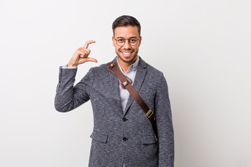 Young business filipino man against a white wall holding something little with forefingers, smiling and confident.