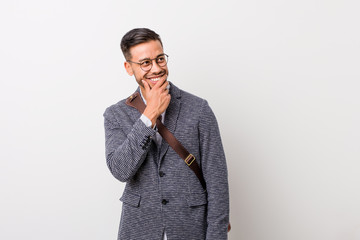 Young business filipino man against a white wall keeps hands under chin, is looking happily aside.