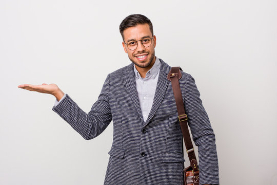 Young Business Filipino Man Against A White Wall Showing A Copy Space On A Palm And Holding Another Hand On Waist.