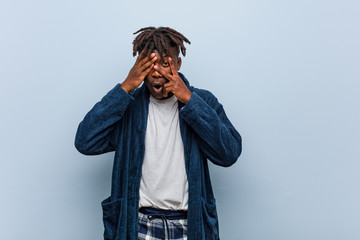 Young african black man wearing pajama blink through fingers frightened and nervous.