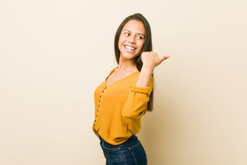 Young hispanic woman against a beige background points with thumb finger away, laughing and carefree.