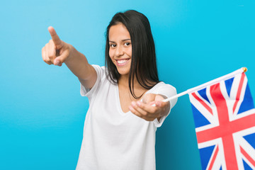 Young hispanic woman holding a united kingdom flag cheerful smiles pointing to front.